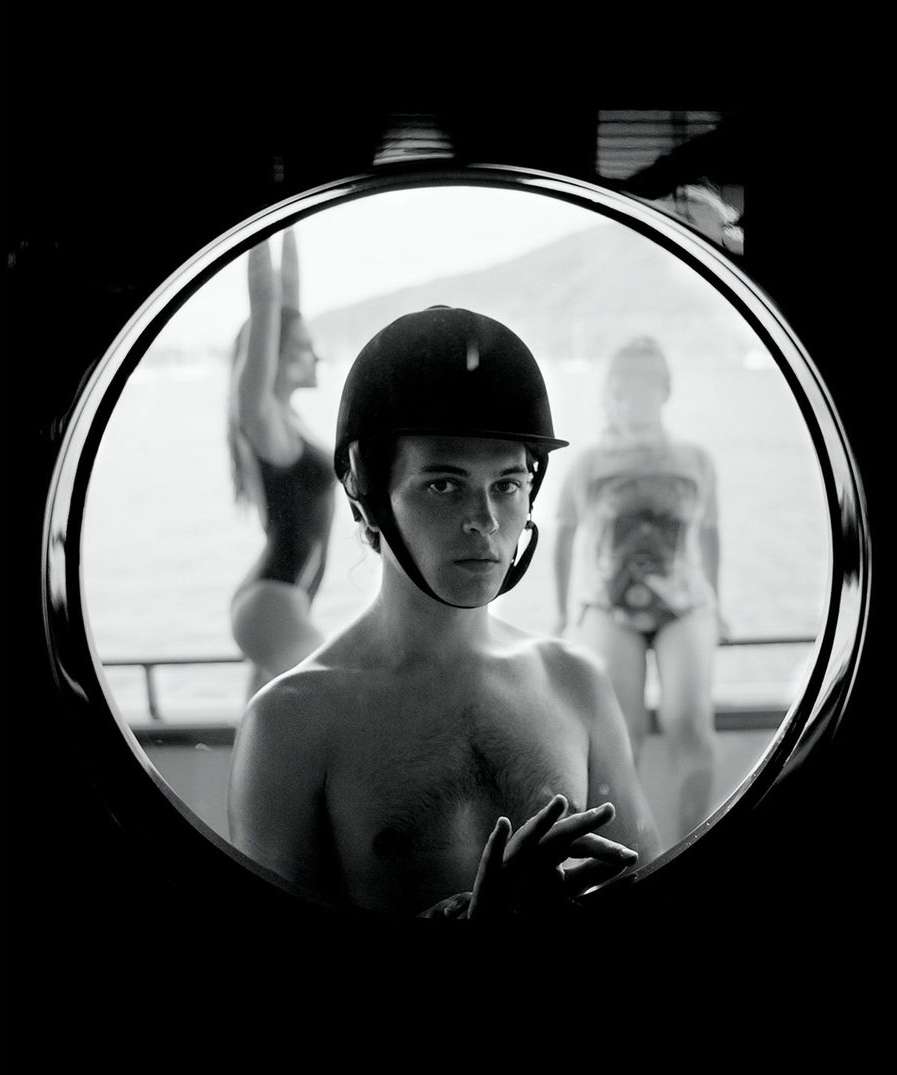 Black and white portrait through circular window of shirtless man in riding helmet with two women in swimsuits in soft focus behind.