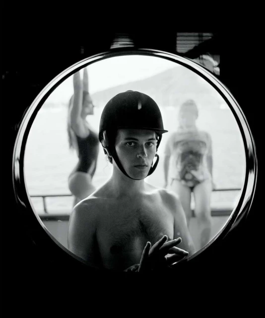 Black and white portrait through circular window of shirtless man in riding helmet with two women in swimsuits in soft focus behind.
