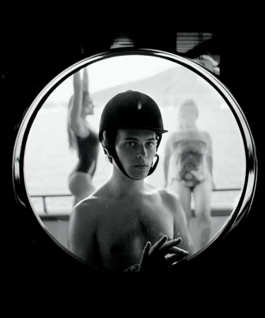 Black and white portrait through circular window of shirtless man in riding helmet with two women in swimsuits in soft focus behind.