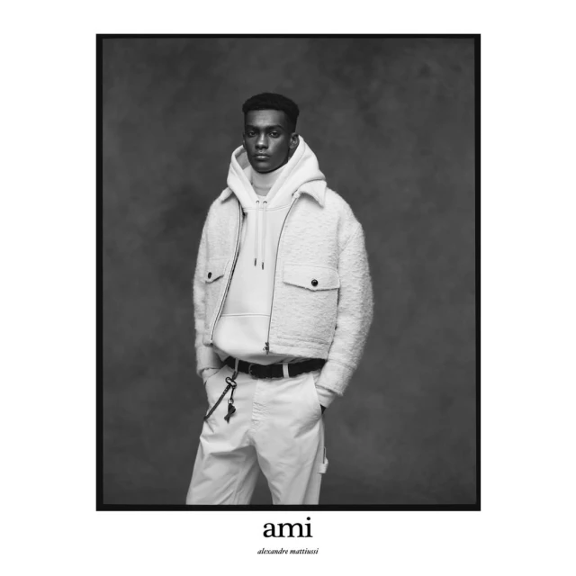 Black and white portrait of a male model in a white hooded sweatshirt and white jacket, posing in front of a dark gray background.