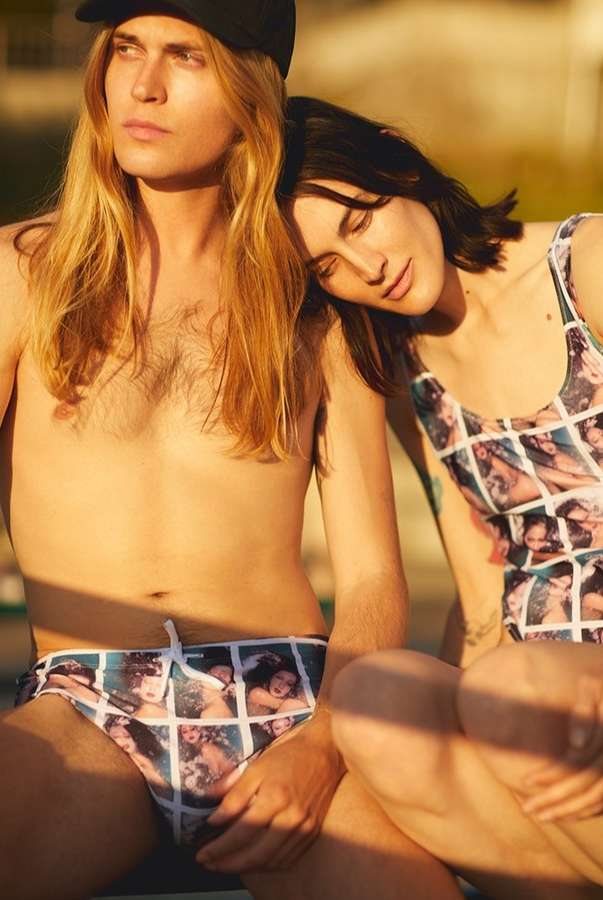A stylish couple poses outdoors in matching swimwear featuring a unique grid pattern with face portraits. Warm, natural light enhances the scene.