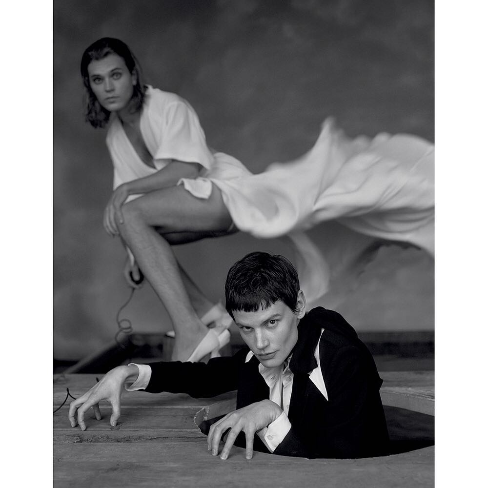 Monochrome studio portrait of two young adult fashion models, one partially emerging from the floor, both in unique poses and attire.