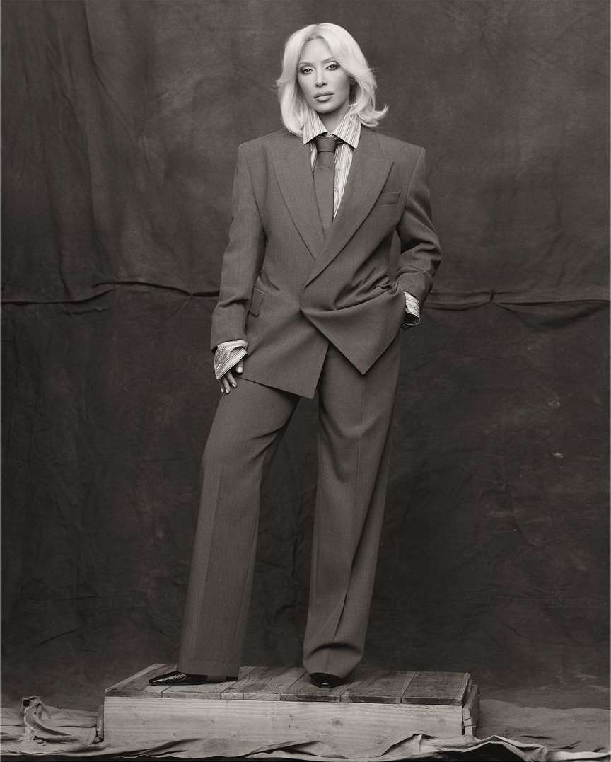 Woman poses in a grey suit and tie, standing on a wooden crate against a neutral backdrop in a studio setting; full length shot.