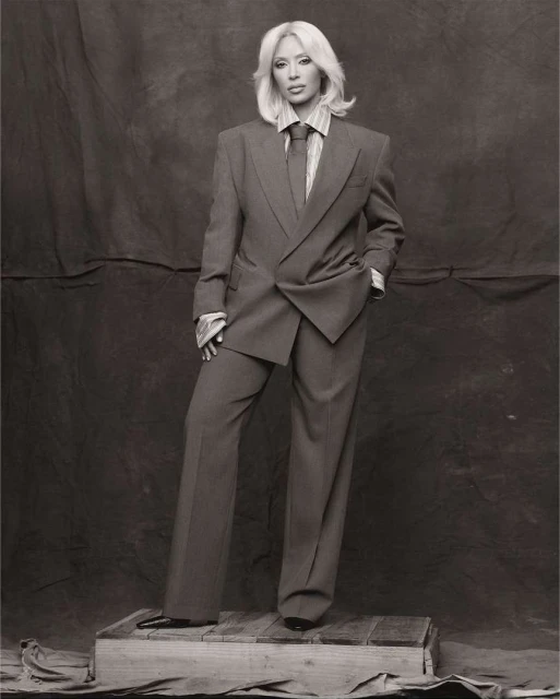 Woman poses in a grey suit and tie, standing on a wooden crate against a neutral backdrop in a studio setting; full length shot.