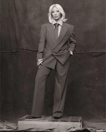 Woman poses in a grey suit and tie, standing on a wooden crate against a neutral backdrop in a studio setting; full length shot.