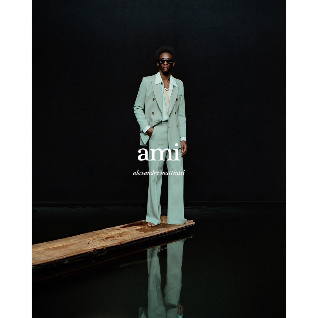 A man in a mint green suit stands confidently on a wooden plank reflecting in water against a black background in a studio shot.