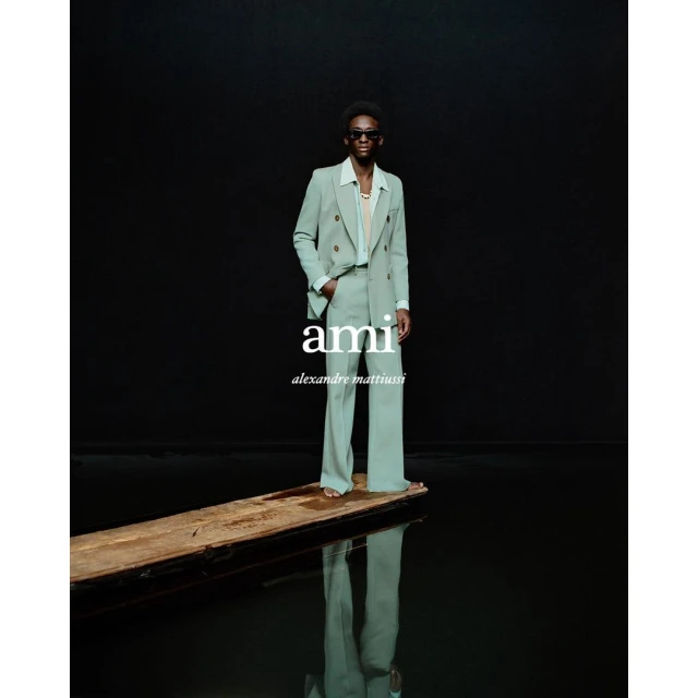 A man in a mint green suit stands confidently on a wooden plank reflecting in water against a black background in a studio shot.