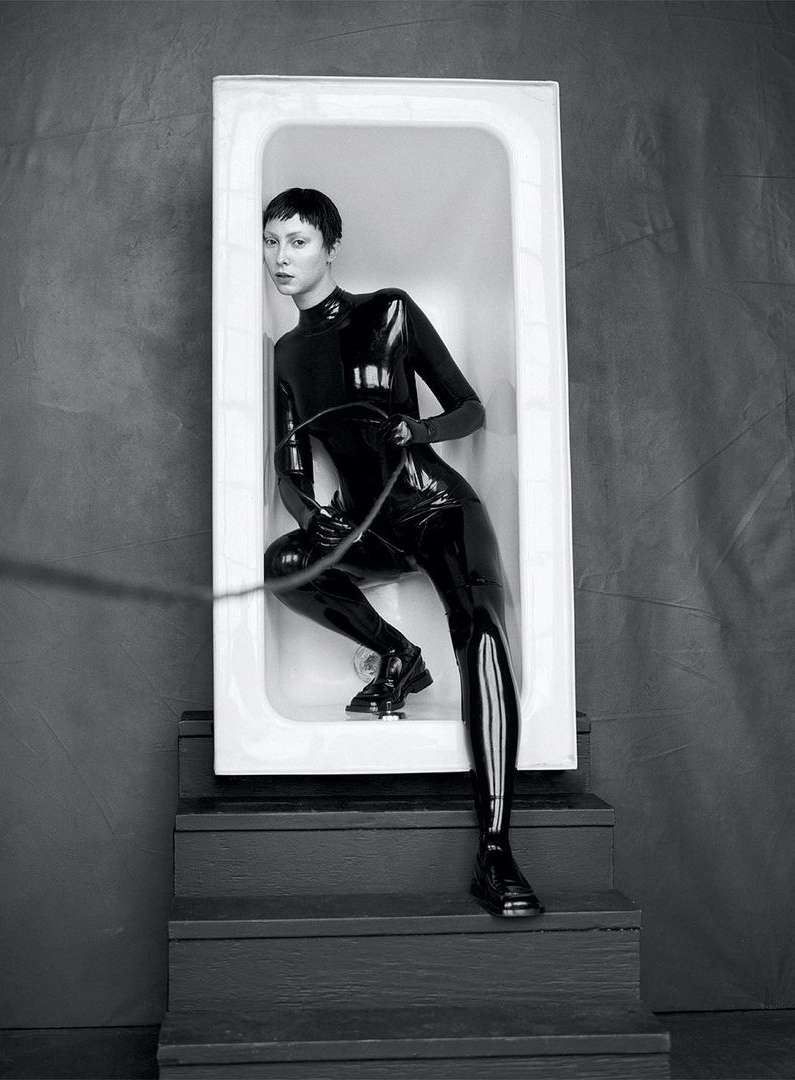 Monochrome portrait of a woman in a shiny black bodysuit, inside a white bathtub on stairs, holding a whip-like object.