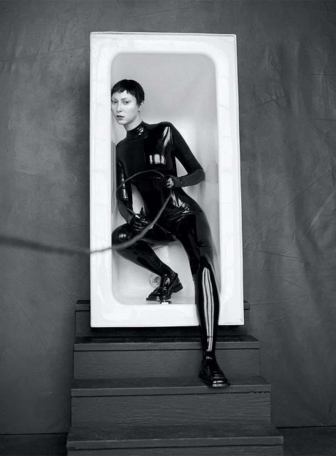 Monochrome portrait of a woman in a shiny black bodysuit, inside a white bathtub on stairs, holding a whip-like object.