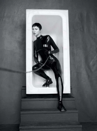 Monochrome portrait of a woman in a shiny black bodysuit, inside a white bathtub on stairs, holding a whip-like object.
