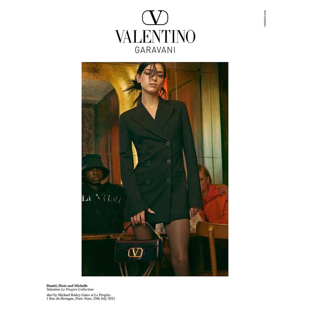 Fashion editorial shot of an adult female in a black blazer dress holding a Valentino purse, posing indoors with other people in the background.