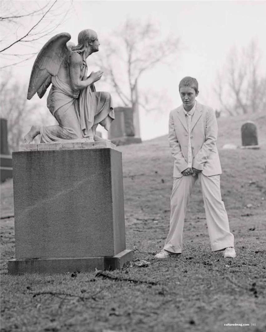 Young adult poses in suit beside angel statue in graveyard on overcast day, wearing sneakers; monochromatic, sepia toned image.