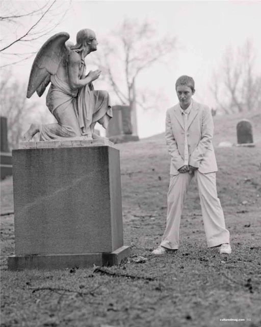 Young adult poses in suit beside angel statue in graveyard on overcast day, wearing sneakers; monochromatic, sepia toned image.