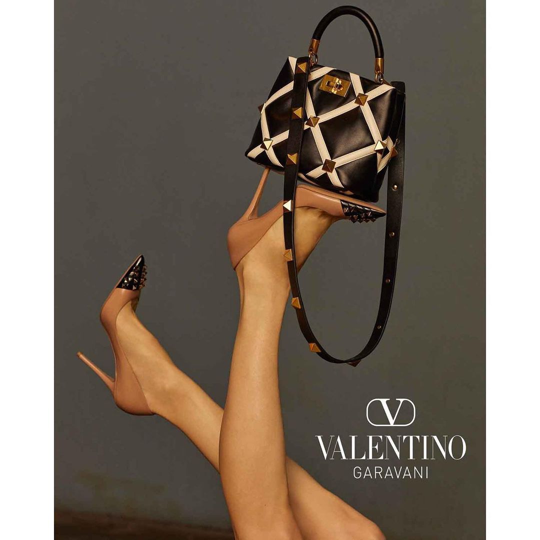 Valentino handbag and studded high heel shoes on a grey backdrop in a studio shot. Fashion advertisement for luxury accessories.