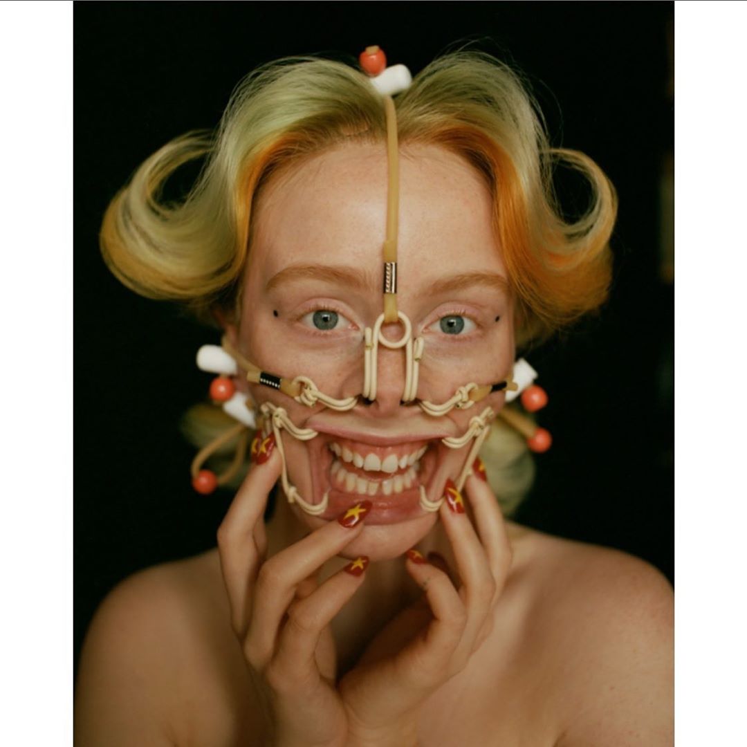 Close-up of a woman with strawberry blonde hair, styled with curls and visible orthopaedic face gear, posing in front of a black background.