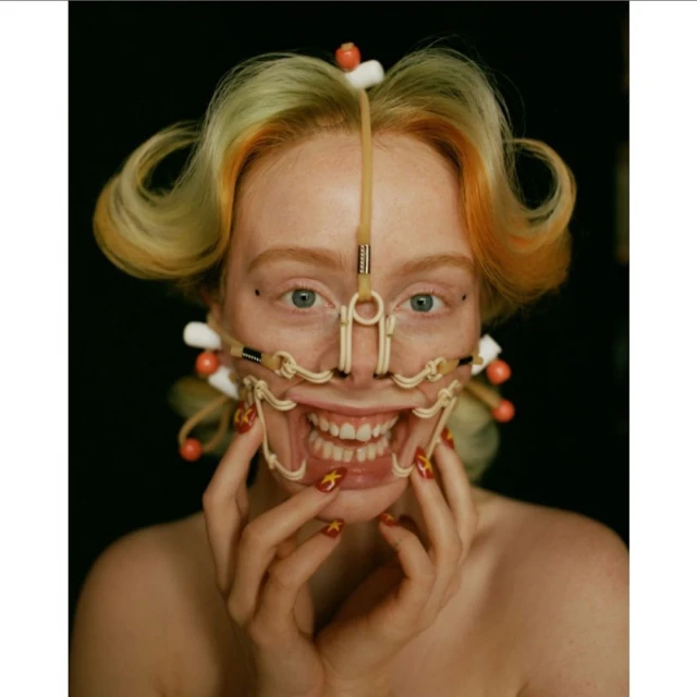 Close-up of a woman with strawberry blonde hair, styled with curls and visible orthopaedic face gear, posing in front of a black background.
