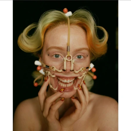 Close-up of a woman with strawberry blonde hair, styled with curls and visible orthopaedic face gear, posing in front of a black background.