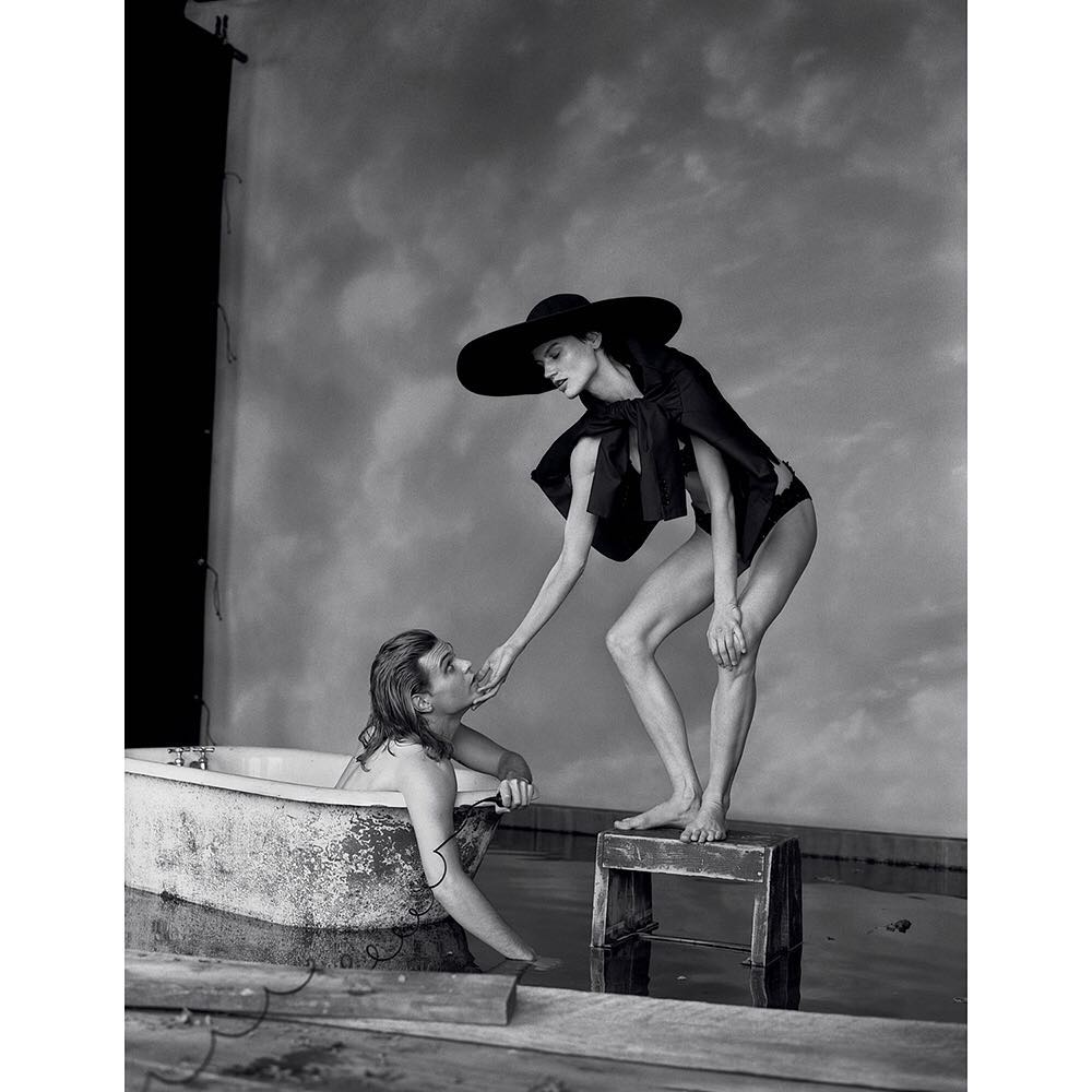 Black and white fashion shot of a woman in a black hat standing on a stool and holding the face of a man in a bathtub.