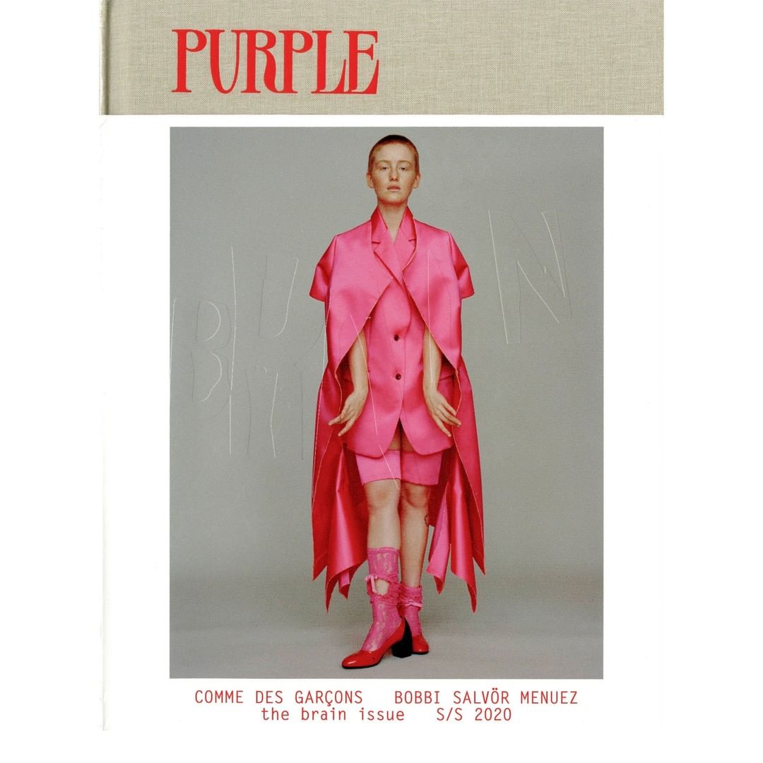 Fashion editorial shot of a person in a pink outfit with a book cover with the word PURPLE in red against a grey background.