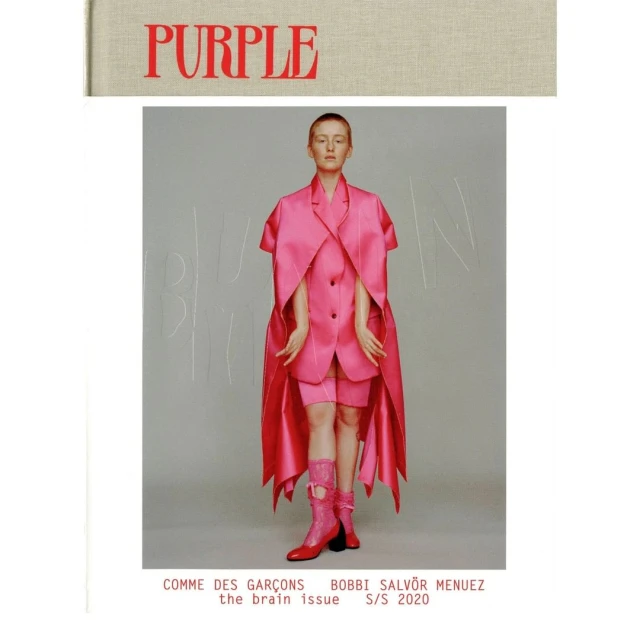 Fashion editorial shot of a person in a pink outfit with a book cover with the word PURPLE in red against a grey background.