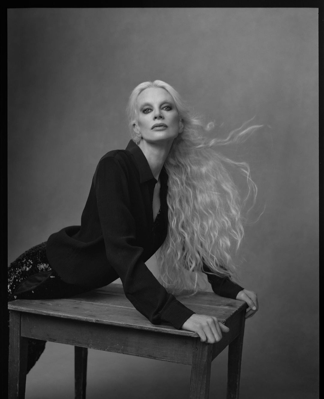 Monochrome portrait of a woman with long blonde wavy hair, wearing a dark jacket, posing on a wooden table in a photo studio.
