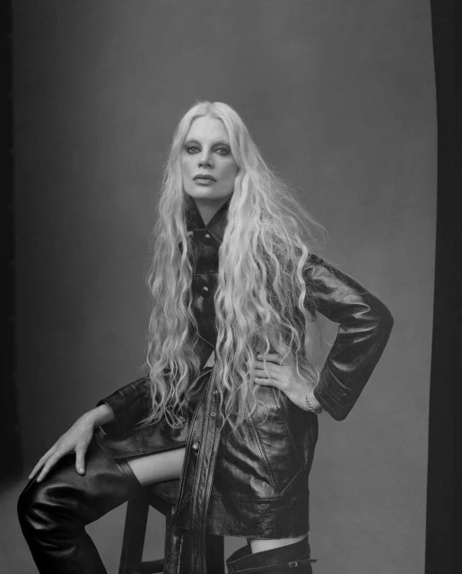Monochrome portrait of a woman with long wavy blonde hair sitting in a black leather jacket and thigh-high boots in a studio setting.