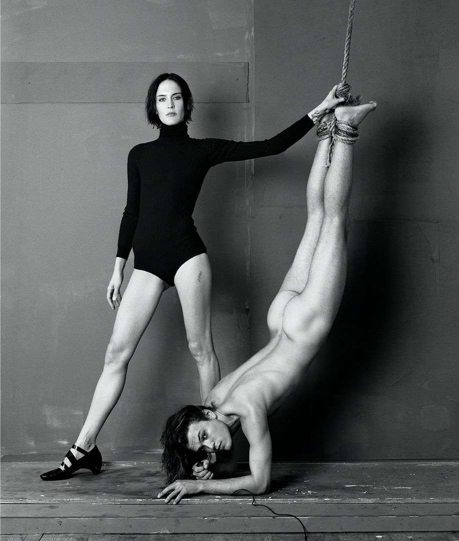 Black and white studio shot: woman stands in black bodysuit, one arm on a partially nude person suspended upside down by a rope.