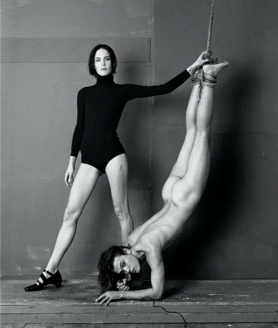 Black and white studio shot: woman stands in black bodysuit, one arm on a partially nude person suspended upside down by a rope.