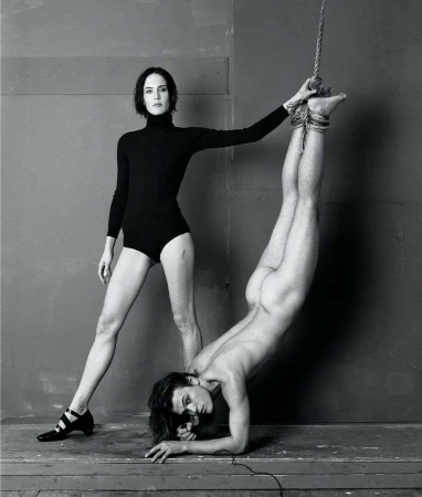 Black and white studio shot: woman stands in black bodysuit, one arm on a partially nude person suspended upside down by a rope.