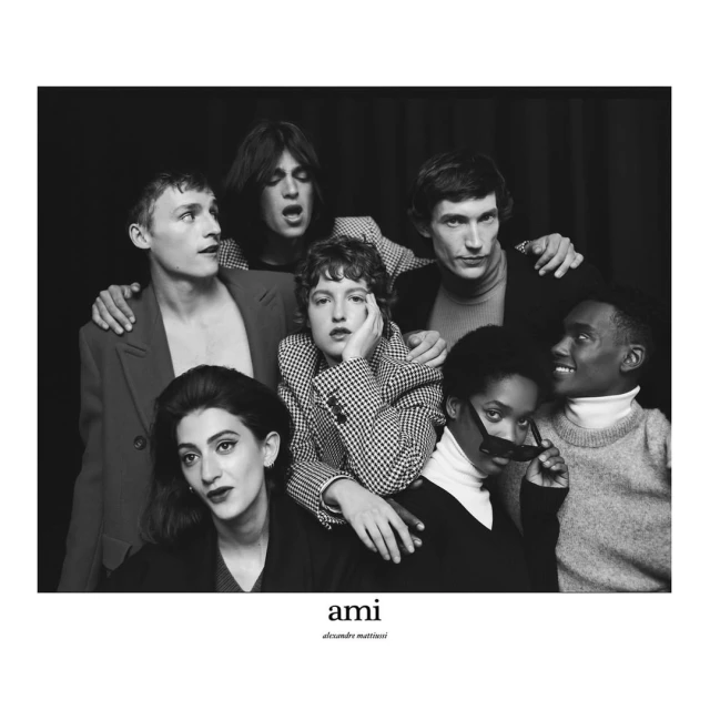 Group portrait of seven people posing in front of a dark background. The group includes men and women in a variety of fashionable clothing.