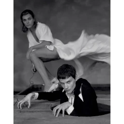 Black and white portrait of two models, one in a white robe and one emerging from a floor opening in a dark suit.