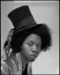 Portrait of a Black man with an afro wearing a top hat with a star patterned scarf and a textured jacket against a plain background.