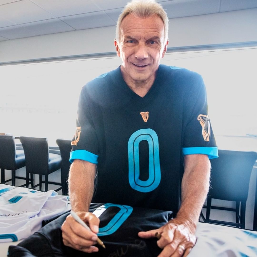 Adult male autographs a black sports jersey with a number '0' in an indoor setting with natural light and chairs visible.