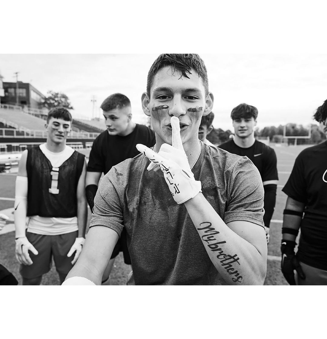 Monochrome image features an adult male footballer gesturing for silence, wearing eye black, gloves, and a T-shirt, standing on a football field.