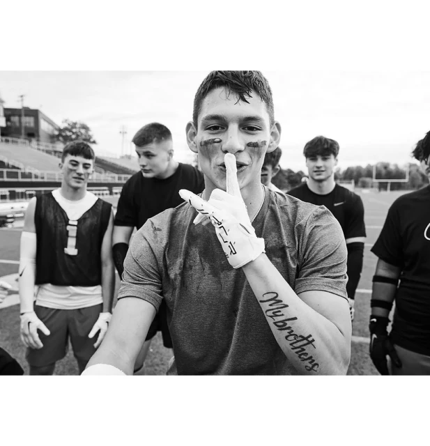 Monochrome image features an adult male footballer gesturing for silence, wearing eye black, gloves, and a T-shirt, standing on a football field.