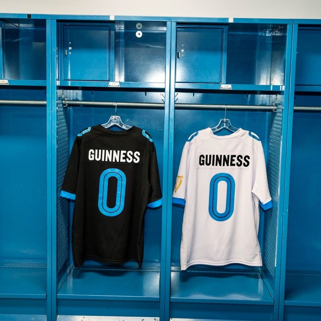 Two Guinness-branded jerseys, one black and one white, hang in adjacent blue metal lockers, presenting a clean and symmetrical locker room scene.