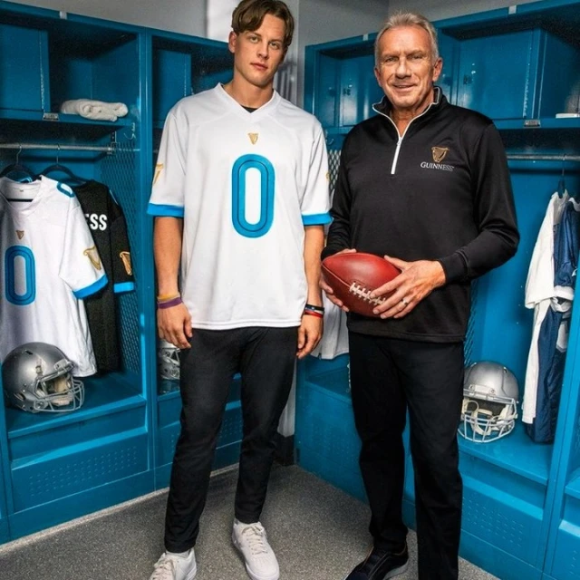 Two adult men pose in a locker room, one holding an American football, wearing Guinness branded apparel and jerseys, with lockers in the background.