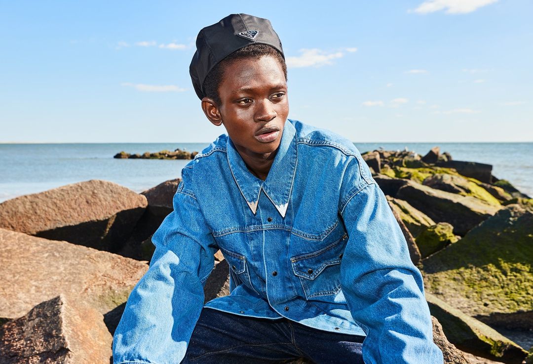 Man in denim jacket and black Prada hat poses on a rocky beach with blue sky and ocean in the background, portrait-style, outdoors.