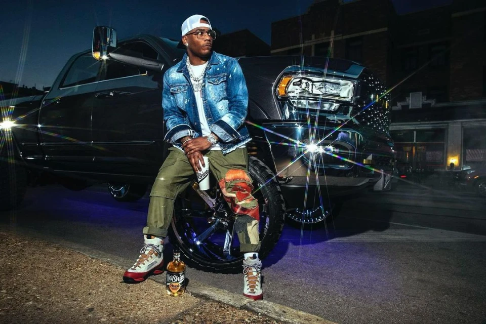 Adult male sitting on a truck tire wearing a denim jacket and sunglasses at night in an urban area with a bottle of liquor.