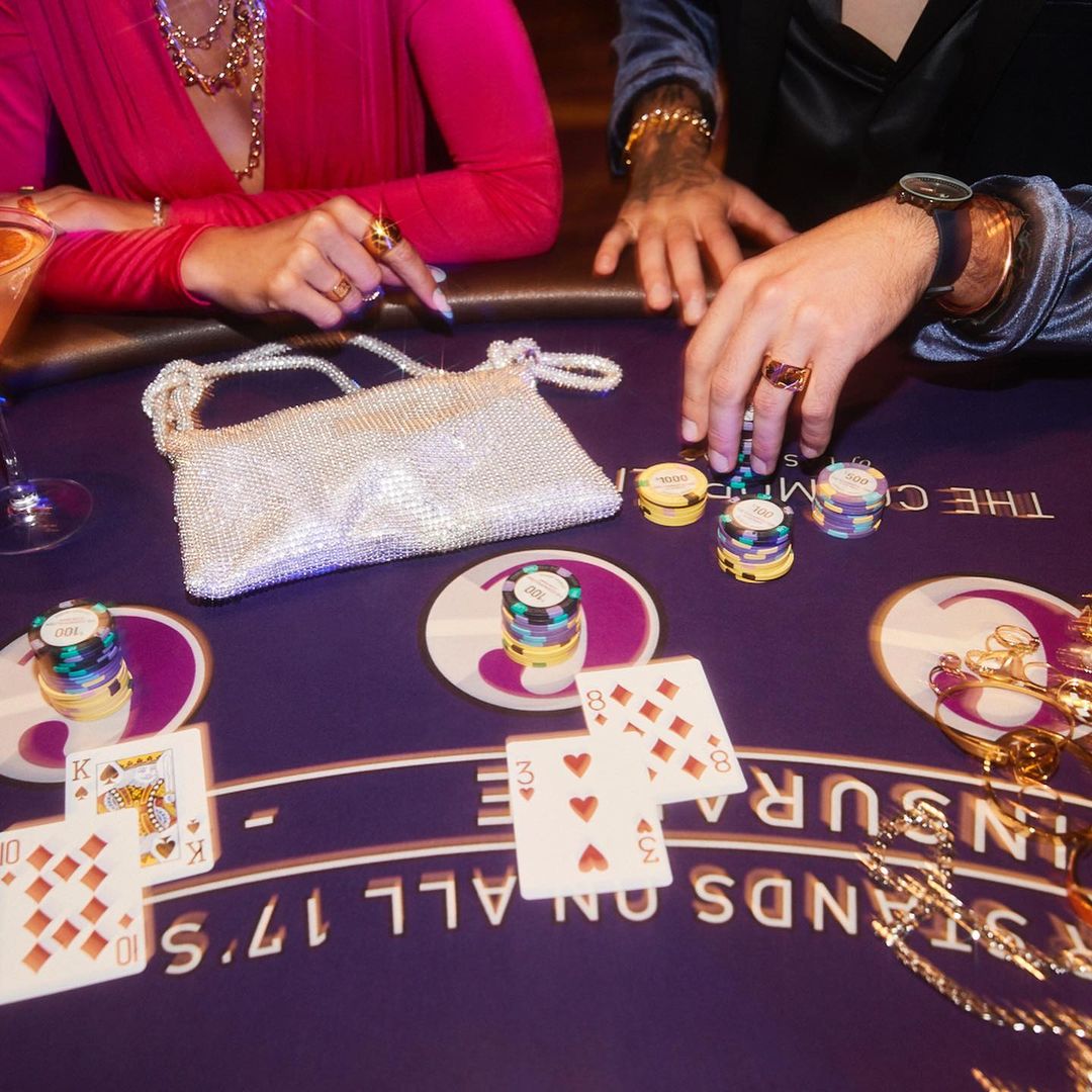 Close-up of a blackjack table with cards, chips, and two people, one in pink and one in blue, with jewelry visible in the frame.