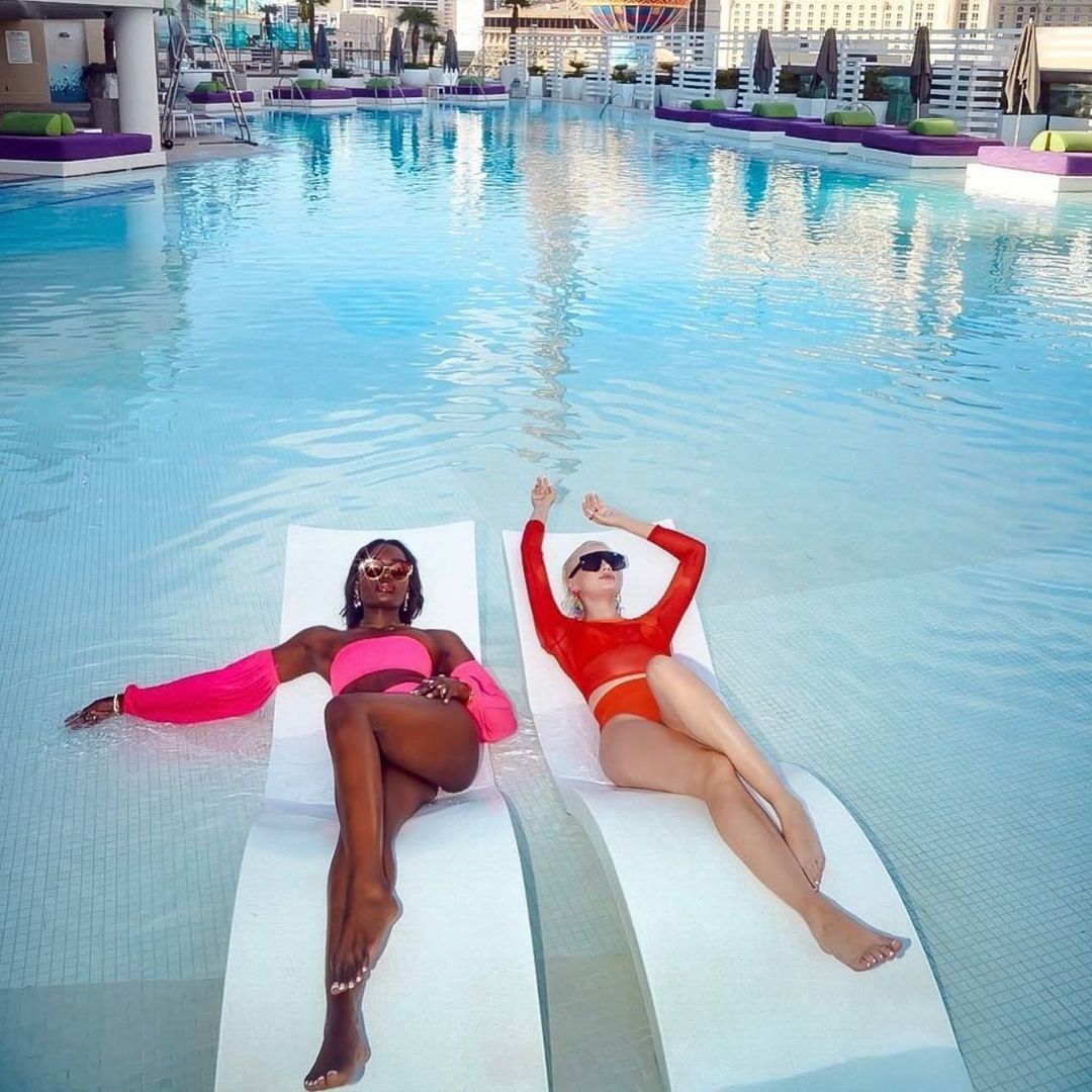 Two women in swimsuits lounge on white chairs in a pool at an upscale resort, with city buildings visible in the background, soaking up the sun.