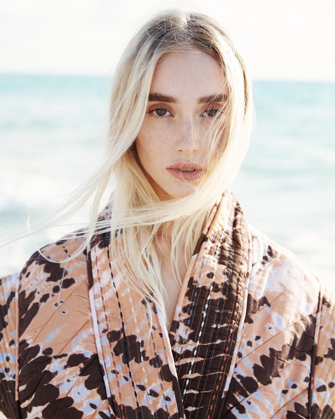 Close-up portrait of a young adult woman with long blonde hair, wearing a patterned coat, posing outdoors with a blurred beach background.