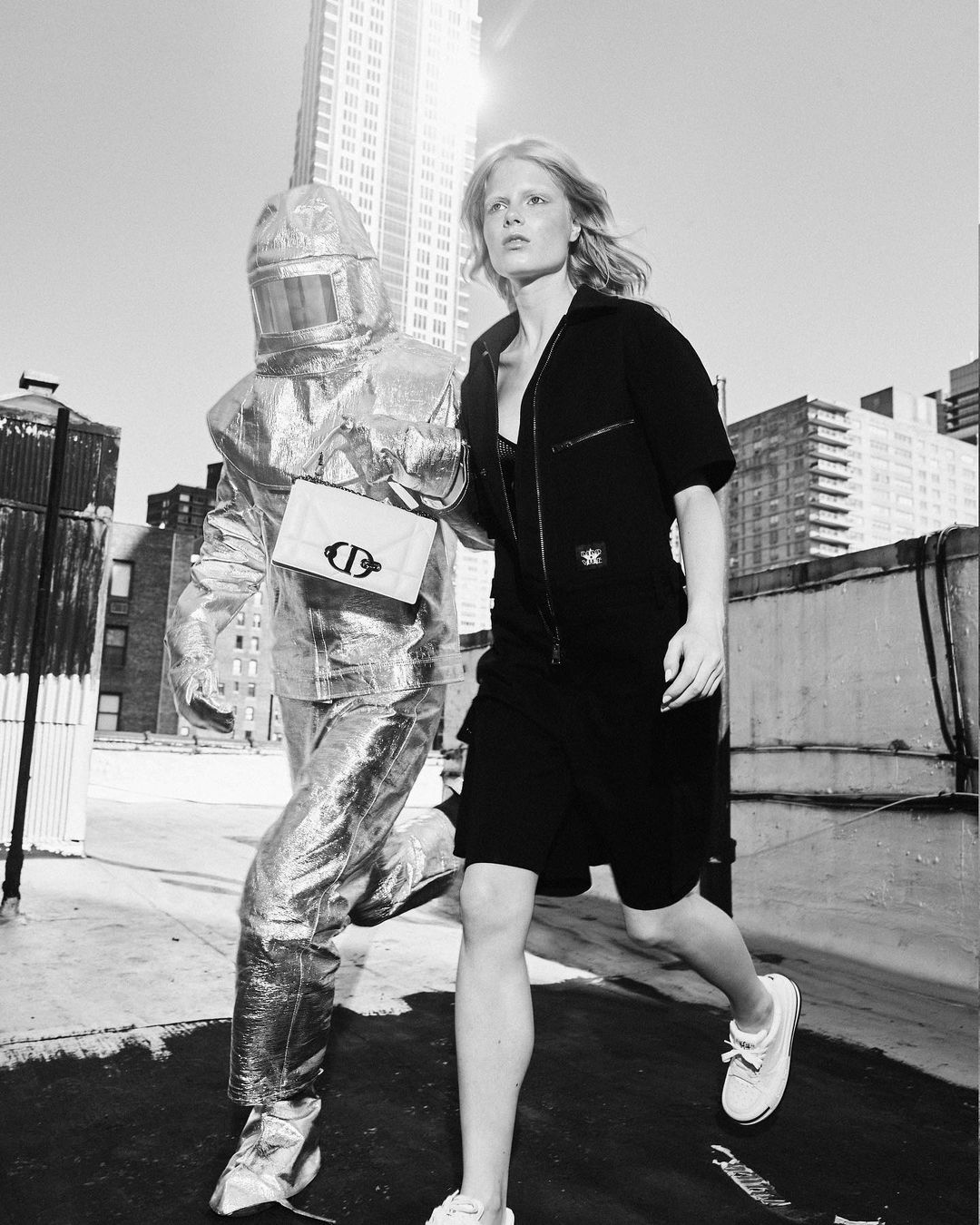 Two models run outdoors on a sunny day, one in a black dress and sneakers and one in a metallic protective suit and carrying a purse.