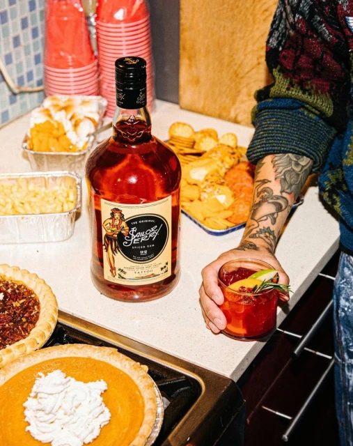 Bottle of Sailor Jerry rum surrounded by pies and snacks on a counter, with a tattooed arm holding a cocktail in a glass.