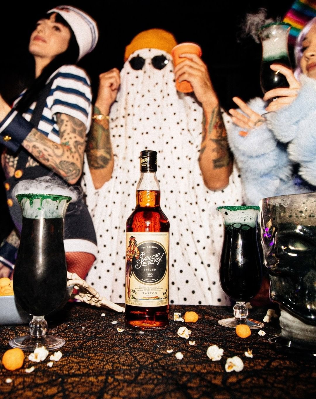 Bottle of Sailor Jerry sits on a table with Halloween cocktails, popcorn, and three partygoers in festive outfits. 