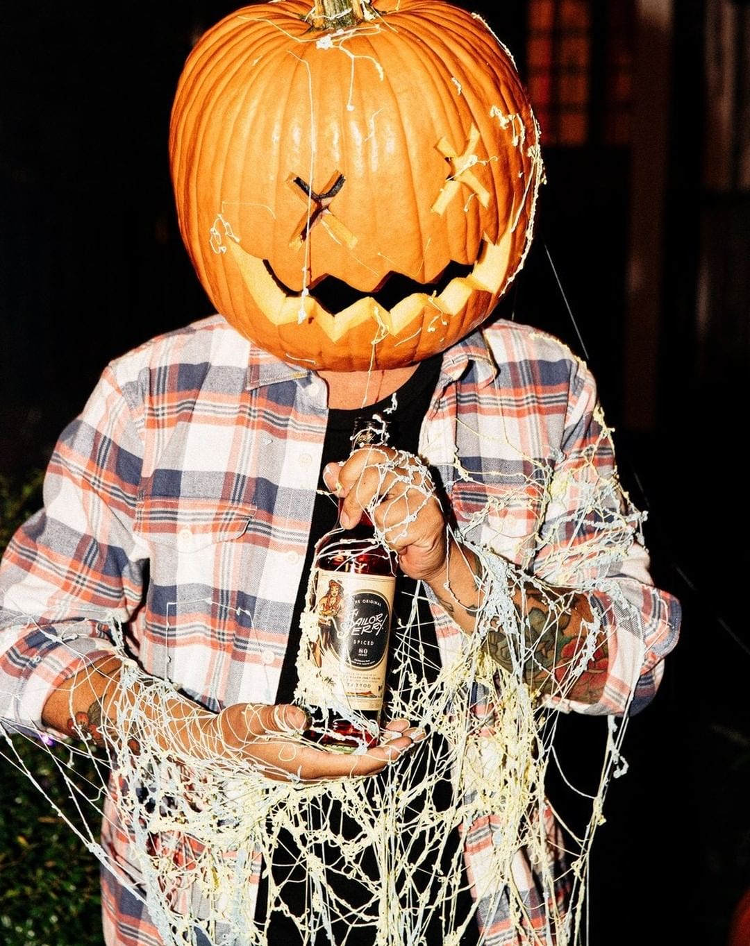 Person with carved pumpkin head holding liquor bottle with cream-colored web accents against a dark background.