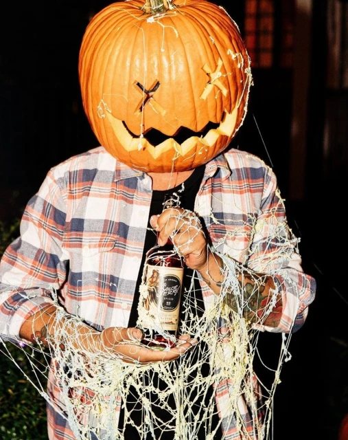 Person with carved pumpkin head holding liquor bottle with cream-colored web accents against a dark background.
