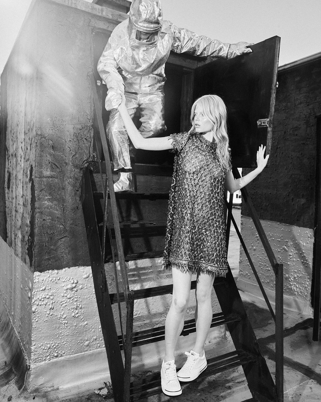 Monochrome shot of a woman in a sequined dress standing on stairs with a man in a protective suit reaching out to her.