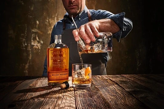 A bartender in an apron pours whiskey from an ice-filled glass into a rocks glass with ice and an orange peel on a rustic wood table.