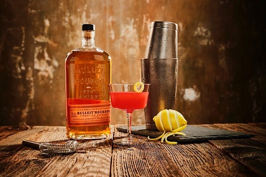 Bulleit Bourbon bottle and a red cocktail in a coupe glass with lemon twist, metal shaker, bar spoon, and lemon on a wooden bar top.